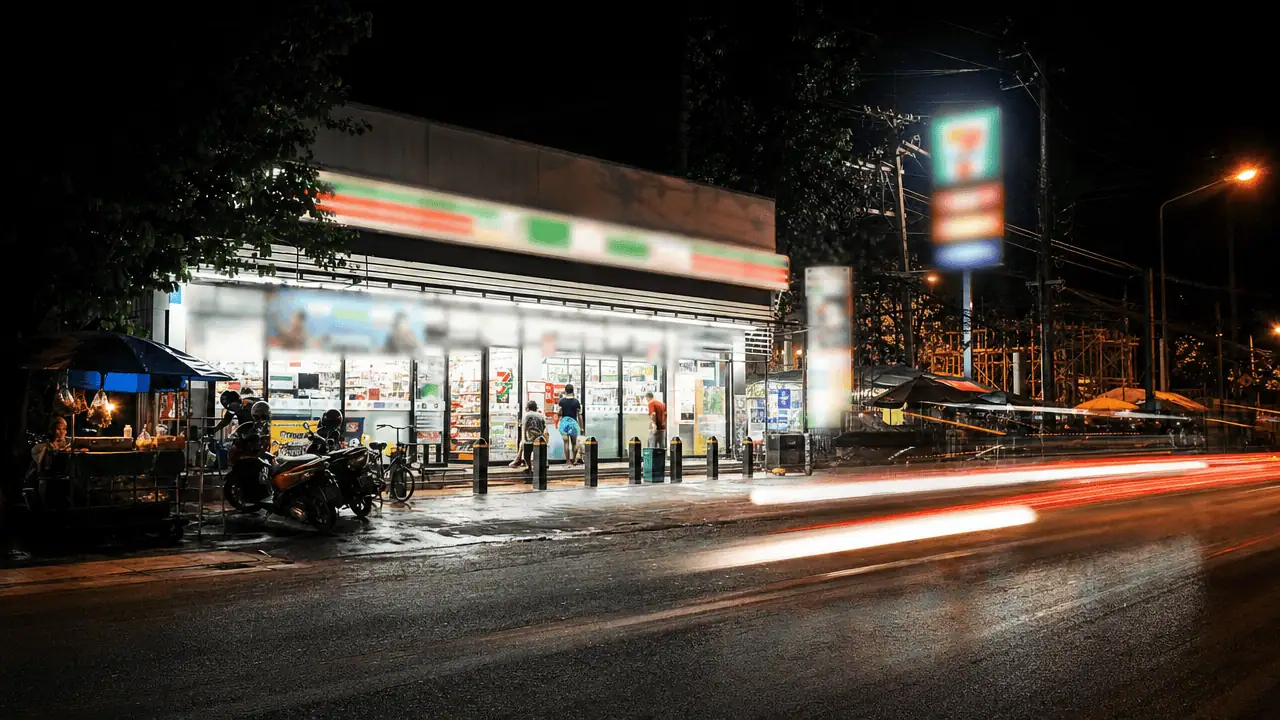เวลาเปิดปิด Google Maps 2026: วิธีตั้ง-แก้ไขให้ตรงจริง 4 ภาพถ่ายบรรยากาศร้านสะดวกซื้อ (ลักษณะคล้าย 7-Eleven) ที่เปิดอยู่ตอนกลางคืน มีลูกค้าเดินเข้าออก รถจักรยานยนต์จอด และมีแสงไฟรถวิ่งผ่านด้านหน้า — เวลาเปิดปิด google maps